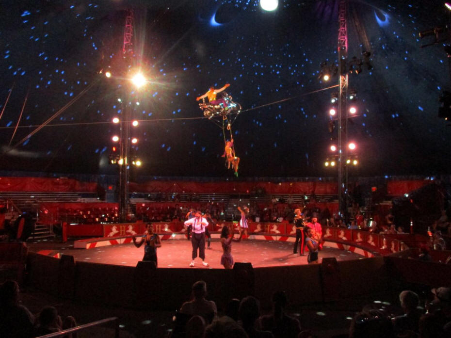 circus preformers riding a bike over a tightrope and otheres below